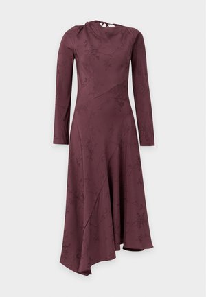 Femme aux cheveux bouclés portant une robe bordeaux à manches longues et col haut avec des motifs floraux et des sandales noires à talons, debout devant un fond uni.