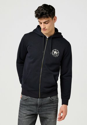 Black zip-up hoodie made of cotton blend, featuring a front logo, kangaroo pockets, and ribbed cuffs, paired with gray jeans.