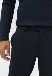 Pantaloni neri con una texture liscia, caratterizzati da un design slim-fit, chiusura con bottone e tasche laterali inclinate. Indossati con una camicia a maniche lunghe scura.
