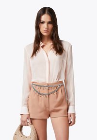Blusa trasparente rosa chiaro abbinata a pantaloni beige a vita alta. Una cintura decorativa in argento con lettere pende in vita. Tiene una borsa beige trapuntata.