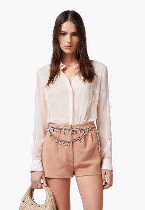 Blusa trasparente rosa chiaro abbinata a pantaloni beige a vita alta. Una cintura decorativa in argento con lettere pende in vita. Tiene una borsa beige trapuntata.