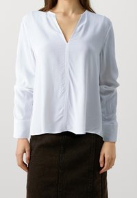 Vit långärmad blus med V-ringning, med en central söm, slät textur och lös passform, tillsammans med en mörk, knälång kjol.