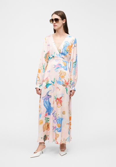 Γυναίκα που φορά μακρύ παστέλ φόρεμα με πολύχρωμα σχέδια θαλάσσιων ζώων, γυαλιά ηλίου, λευκά παπούτσια με χαμηλό τακούνι και βραχιόλι ποδιού, στέκεται σε λευκό φόντο.