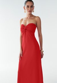 Rode strapless jurk met een gedraaid lijfje, vloerlange ontwerp, gladde stof en zijsplit. Gouden accessoires complementeren de outfit.