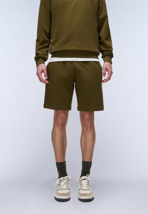 Mann trägt olivgrüne Shorts, passendes Sweatshirt, dunkelgrüne Socken und weiße Sneakers, steht vor einem einfarbigen Hintergrund.