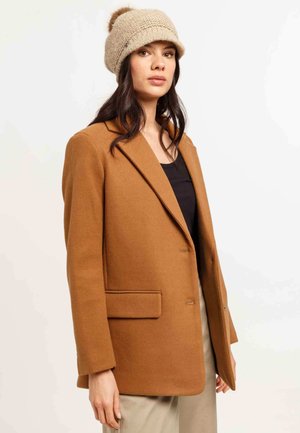 Bruine wolmix blazer met een ingestoken revers, één knoopsluiting en twee voorzakken. Beige gebreide muts met een pompon-detail.