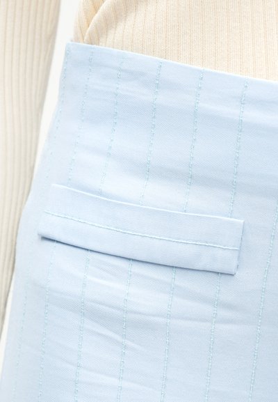Φούστα ανοιχτού γαλάζιου χρώματος με λεπτές ρίγες και οριζόντια τσέπη με καρφίτσα, φορεμένη πάνω από μπεζ πλεκτό τοπ με ρίγες.