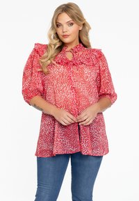 Rode blouse met patroon, een ruches-kraag en een sleutelgatdetail. Doorzichtig materiaal, ballonmouwen en een losse pasvorm. Gepaarde met blauwe jeans.