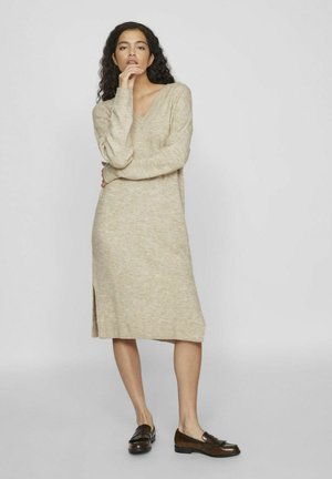 Strikket beige kjole med V-hals og lange ermer, med en sideslitt, kombinert med mørkebrune loafers. Myk tekstur og avslappet passform.