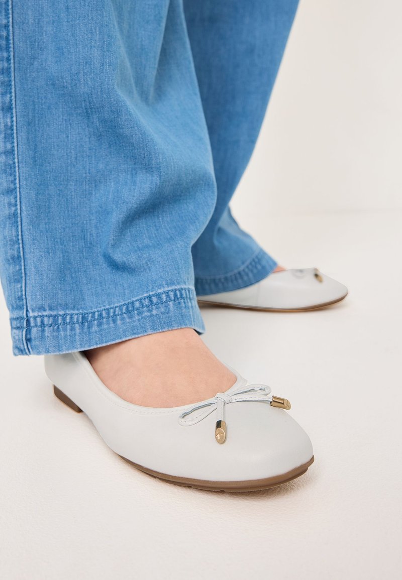 Ljust grå ballerina-skor med rund tå, dekorativ rosett och gyllene accenter. Slätt lädermaterial, kombinerat med blå denimbyxor.
