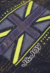 Zwarte stof met een groot geel en grijs patroon dat op een vlag lijkt, met de tekst "J&JOY" in het wit en "UNITED KINGDOM" in het geel.