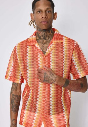 Homme avec des tatouages au cou et au bras porte une chemise à manches courtes à motifs orange, jaune et blanc boutonnée, de face sur un fond uni.