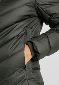 Veste gonflée vert foncé avec motif matelassé diagonal. Comprend un poignet côtelé, une poche latérale et une texture extérieure lisse et brillante.