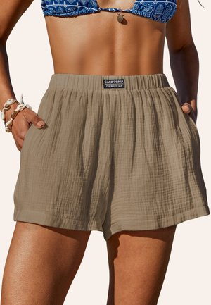 Beige shorts in lightweight, textured fabric with a loose fit and elastic waistband. Notable design includes front pockets and brand label.