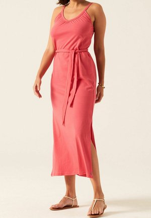 Femme portant une robe maxi rose corail sans manches avec une ceinture à la taille et des sandales plates beige, debout devant un fond neutre.