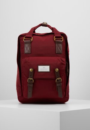 Roter rechteckiger Rucksack mit Fronttasche, braunen Lederriemen mit Messingschnallen und einem oberen Griff, stehend auf einer weißen Oberfläche.