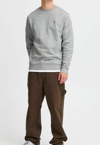 Sudadera gris con cuello redondo y puños acanalados, con un pequeño diseño bordado en el pecho. Pantalones cargo marrones con bolsillos laterales.
