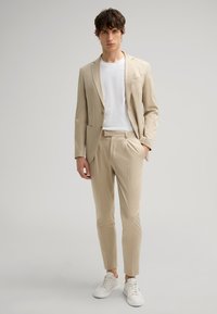 Chaqueta de traje beige con pantalones slim-fit a juego, camiseta blanca debajo y zapatillas blancas. Tejido suave, diseño ajustado, sin patrones.