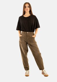 Pantaloni in velluto a coste marrone con grandi tasche frontali, abbinati a una maglietta nera a trama morbida e ampia e sneakers beige con dettagli bordeaux.