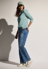 Sudadera de manga larga azul claro, jeans de mezclilla de pata ancha en azul y zapatillas off-white con acentos verdes, combinadas con una gorra a juego.