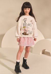 Sweat-shirt beige clair avec broderie florale et texte, associé à une jupe en sequins rose pastel. Bottines noires avec détail boucle.