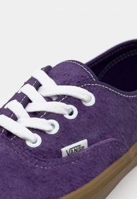 Lila mockaskor med vita snören och sömmar. Har en tan gummisula och en liten vit "VANS"-etikett på sidan.