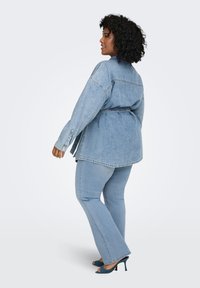 Γυναίκα με σγουρά μαλλιά που φοράει τζιν μπουφάν με ζώνη, ανοιχτόχρωμα μπλε τζιν και μπλε ψηλοτάκουνα σανδάλια, στέκεται και κοιτάει στο πλάι.