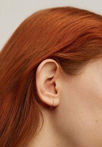 Pendiente de aro dorado con un diseño retorcido colgando de la lóbulo de la oreja, exhibido contra un cabello lacio de color castaño rojizo y un fondo neutro.