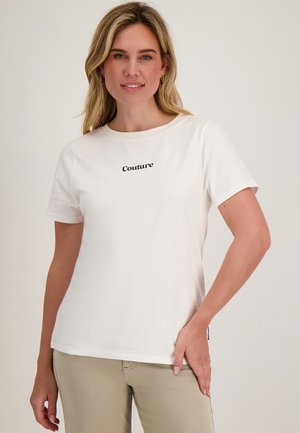 Blonde vrouw die een wit shirt met korte mouwen draagt met de tekst "Couture", beige broek, staand tegen een effen lichte achtergrond.