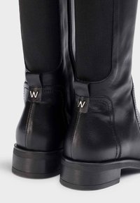 Bottes noires en cuir, arrivant au genou, avec des côtés élastiques, présentant un petit logo argenté et un épais talon noir robuste.