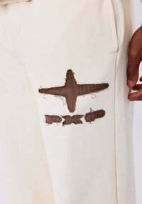 Crème kleurige broek met bruine geborduurde ster en tekstdetails. Materiaal is zacht met een licht getextureerde afwerking. Zichtbare stikselaccenten.