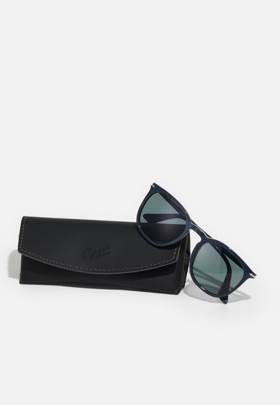 Temno mornarske sončne očala z zelenimi lečami in srebrnimi detajli, ki počivajo ob črni usnjeni etui z embosiranim blagovno znamko in kontrastnim šivom.