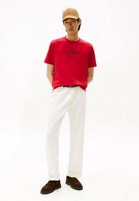 Rød bomulds T-shirt med "Tommy Hilfiger" logo, parret med hvide bukser og brune ruskinds sko. En simpel kasket fuldender outfittet.