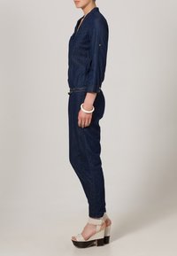 Denim jumpsuit i mörkblått, med v-ringning, långa ärmar och midjebälte. Bärs med vita platåskor och ett armbandsur.