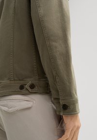 Giacca in denim verde oliva con taglio dritto. Caratterizzata da chiusura a bottone ai polsini e orlo regolabile a due bottoni. Tessuto testurizzato.