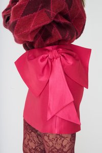 Krótka różowa sukienka z dużą satynową kokardą z tyłu. Dzianinowy sweter w kolorze ciemnoczerwonym z diamentowym wzorem. Modelka ma na sobie rajstopy z koronkowym wzorem.