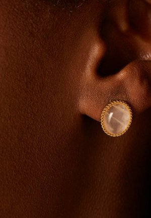 Close-up van een donker gekleurde oor met een ovale, doorschijnende witte edelsteenoorbel met een gouden kralenrand.