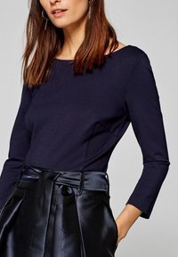 Kvinna klädd i en figursydd mörkblå långärmad topp stoppad i högmidjade svarta satinbyxor med ett brett knytbälte.