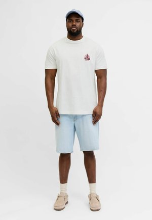 Mann steht und trägt ein weißes T-Shirt mit Grafik, helle Jeansshorts, beige Schuhe mit Socken und eine hellblaue Baseballkappe vor einfarbigem Hintergrund.