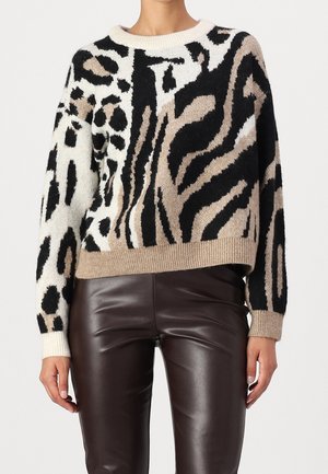 Kobieta ubrana w beżowy, czarny i biały sweter w zwierzęcy wzór, zestawiony z ciemnobrązowymi skórzanymi spodniami na jednolitym tle.
