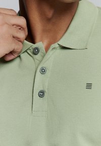 Lichtgroene polo met een gestructureerde stof, drie grijze knopen bij de kraag en een zwart gestreept logo op de linkerkant van de borst.