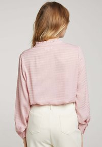Blouse rose clair avec un motif rayé texturé, col volanté et manches longues. Portée avec un pantalon couleur crème, mettant en valeur une coupe décontractée.