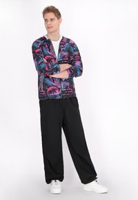 Jachetă cu glugă, cu un model tropical vibrant în roz și albastru, purtată cu pantaloni largi negri și adidași albi.