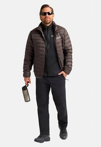 Brown puffer jacket, black zip-up layer underneath, black pants, and black hiking boots. Holding a gray water bottle. Sunglasses worn.