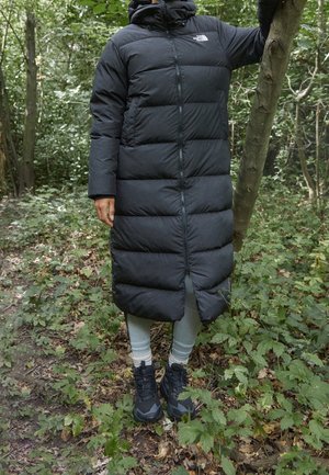 Personne portant un long manteau doudoune noir, des leggings gris clair et des bottes noires, debout dans une forêt verte, tenant un tronc d’arbre d’une main.