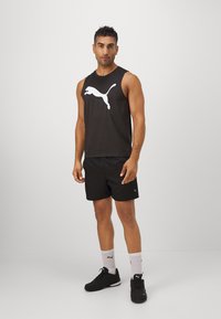 Zwart mouwloos sportshirt met een wit Puma-logo, gecombineerd met zwarte shorts. Accessoires omvatten witte sokken en zwarte sneakers.