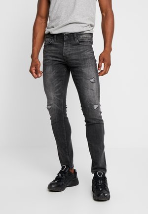 Man draagt slanke zwarte jeans met distressed details, een grijs T-shirt en zwarte sneakers, staand tegen een effen lichte achtergrond.