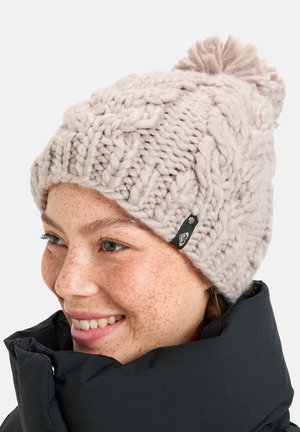 Lichtbeige gebreide muts met dikke kabels, afgewerkt met een pompon en een klein logo-label aan de zijkant. Zacht, gestructureerd garen.