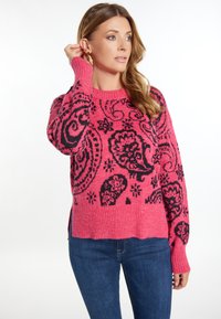 Różowy sweter dziergany z czarnym wzorem paisley, z prążkowanymi mankietami i dołem, z bocznymi rozcięciami dla dodatkowego efektu. Połączony z niebieskimi dżinsami.