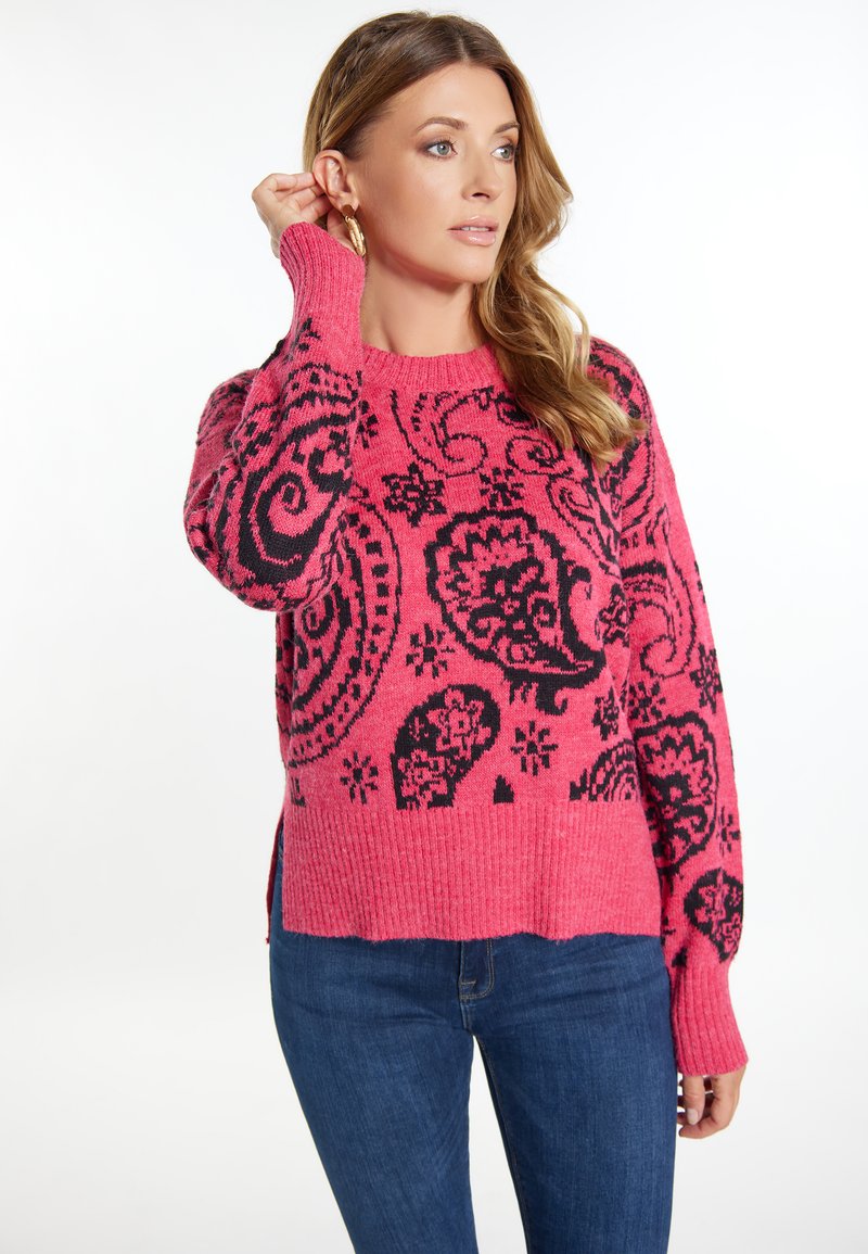 Różowy sweter dziergany z czarnym wzorem paisley, z prążkowanymi mankietami i dołem, z bocznymi rozcięciami dla dodatkowego efektu. Połączony z niebieskimi dżinsami.